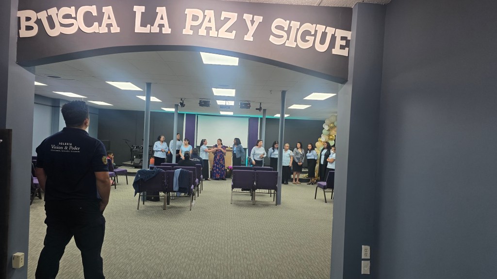 Interior of a meeting space with the sign 'BUSCA LA PAZ Y SIGUE' visible. A group of people stands in the background, some wearing blue shirts, and several chairs are lined up facing the front.